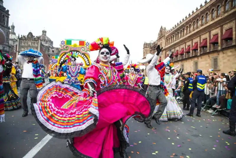 Mulher dançando com um vestido predominantemente rosa, mas com outras cores, a cara totalmente pintada de branco, porém com a região dos olhos todo preto imitando uma caveira. Atrás dela, pessoas com roupas coloridas e sombreiros mexicanos e com o rosto com pintura semelhante desfilam por uma rua da Cidade do México. O conceito básico do Cemitério Mexicano.