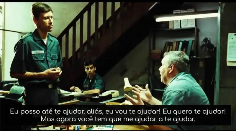 Imagem do filme tropa de Elite. Nela aparecem o Soldado Paulo (homem, branco, magro, cabelo curto penteado castanho) de pé com as mãos entrelaçadas utilizando o uniforme da Polícia Militar do Rio de Janeiro. Sentado com as mãos abertas o Sargento Rocha   (homem, branco, gordo, cabelo curto penteado grisalho). Ao fundo um soldado olhando para o Sargento Rocha (homem, branco, cabelo curto, penteado, preto). Todos estão em uma sala desorganizada com muitos papéis espalhados sobre a mesa e uma estante com pastas de documentos ao fundo e uma escada que demonstra que a sala fica em algum lugar em que as pessoas devem descer para acessá-la.