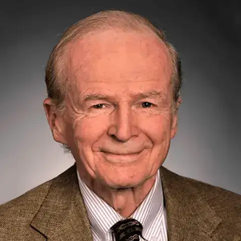 Retrato de John D.C. Little. Um senhor idoso sorridente, de cabelos grisalhos e penteados para trás, usando paletó marrom, camisa listrada e gravata escura, posando diante de um fundo neutro.