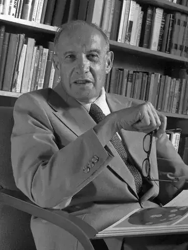 Peter Drucker foto em preto e branco. Homem branco, por volta de 70 anos, trajando terno com gravata e segurando um óculos de leitura frente a um livro com imagens (parece ser um livro infantil). Atrás dele uma estante com vários livros.