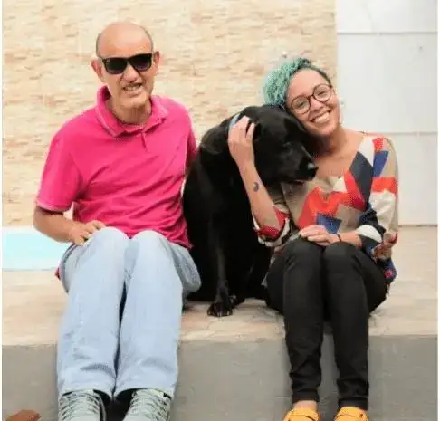 #pracegover Na imagem, está Beto Pereira, Terry (seu cão-guia) e eu. Beto está sorrindo e veste uma camisa rosa e uma calça jeans. Terry está abraçado comigo e é um labrador de cor preta. Eu estou sorrindo e visto uma blusa estampada e uma calça preta, e meu cabelo está verde.
